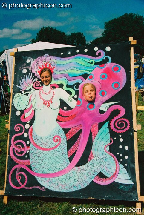 Two girls poke their heads through a mermaid painted board at Big Green Gathering 2003. Cheddar, Great Britain. © 2003 Photographicon