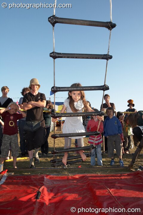 A young girl attempts the challenge of climbing a free-rotating rope ladder at Big Green Gathering 2007. Burrington, Cheddar, Great Britain. © 2007 Photographicon
