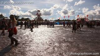 The bright sun reflects off the brown wet muddy expanse silhouetting people walking by at Glade Festival 2007. Aldermaston, Great Britain. © 2007 Photographicon