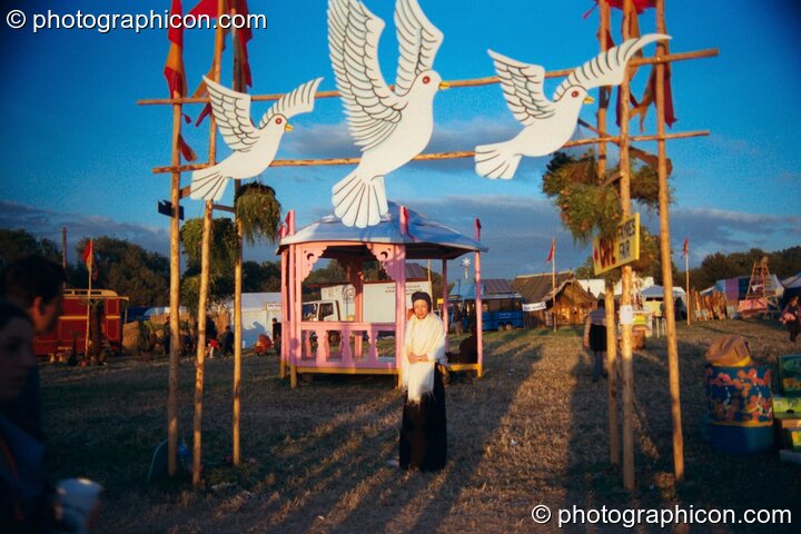 A woman stands under a doves-of-peace archway bathed in the setting sun at Glastonbury Festival 2002. Pilton, Great Britain. © 2002 Photographicon