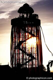 Silhouetted against the setting sun, a tall decorated scaffold tower with viewing platform rises above The Park at Glastonbury Festival 2008. Pilton, Great Britain. © 2008 Photographicon