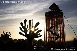 Silhouetted against the setting sun, a tall decorated scaffold tower with viewing platform rises above The Park next to cut-out sculptures of organic shapes at Glastonbury Festival 2008. Pilton, Great Britain. © 2008 Photographicon