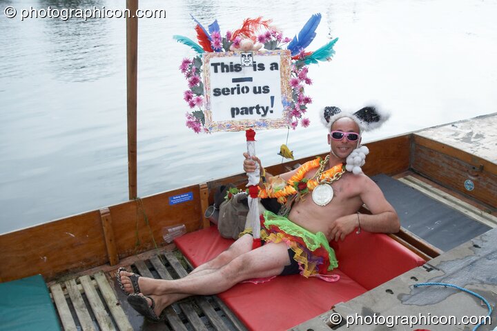 A man in wonky clothes holds a banner while reclining in a boat at the Secret Garden Party 2008. Huntingdon, Great Britain. © 2008 Photographicon