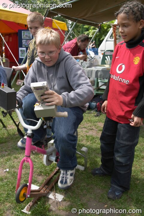 A boy pedals a small bicycle generator so that he can play the Game Boy it powers at Kingston Green Fair 2004. Kingston Upon Thames, Great Britain. © 2004 Photographicon