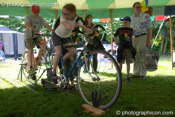 A couple of kids watched by their parents pedal a bicycle generator at Kingston Green Fair 2004. Kingston Upon Thames, Great Britain. © 2004 Photographicon