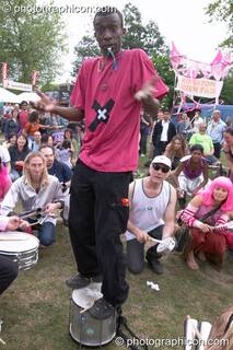 The leader of Rhythms of Resistance stands on his drum and shrugs at Kingston Green Fair 2004. Kingston Upon Thames, Great Britain. © 2004 Photographicon