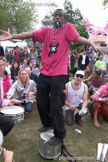 The leader of Rhythms of Resistance stands on his drum and shrugs at Kingston Green Fair 2004. Kingston Upon Thames, Great Britain. © 2004 Photographicon