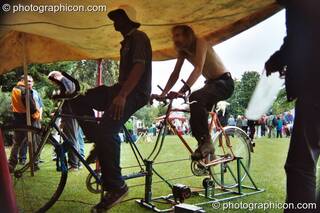 Two mwn on a bicycle generator providing power to a small music stage at Kingston Green Fair 2002. Kingston upon Thames, Great Britain. © 2002 Photographicon