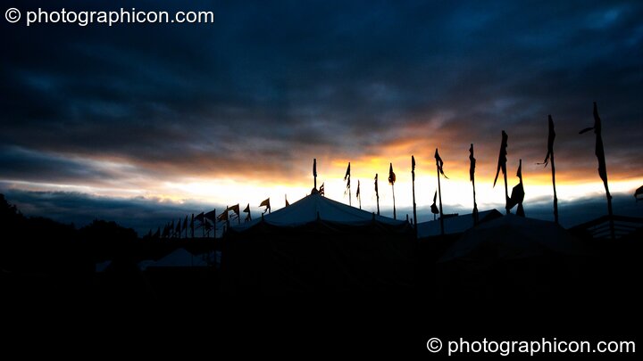 The dawn sun seeps through a crack in ominous black clouds, silhouetting tent and flags at Glastonbury Festival 2005. Pilton, Great Britain. © 2005 Photographicon