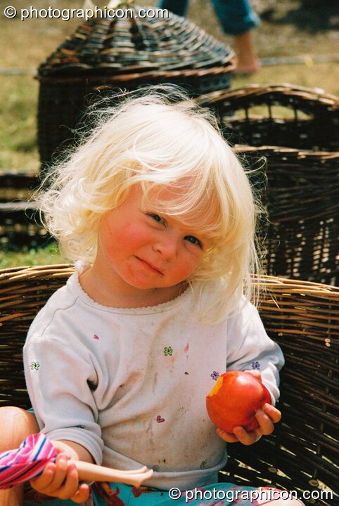 A little girl sits in a basket eating apple at Big Green Gathering 2003. Cheddar, United Kingdom. © 2003 Photographicon