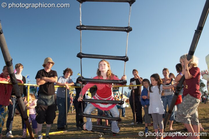 A young girl attempts the challenge of climbing a free-rotating rope ladder at Big Green Gathering 2007. Burrington, Cheddar, United Kingdom. © 2007 Photographicon