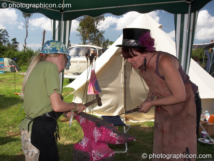 A lady blacksmith holds hot metal while her daughter hammers it into shape at Big Green Gathering 2007. Burrington, Cheddar, United Kingdom. © 2007 Photographicon