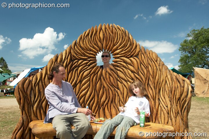 A woman looks on while a man and girl eat lunch seated on a beautifully carved woodedn bench at Big Green Gathering 2007. Burrington, Cheddar, United Kingdom. © 2007 Photographicon