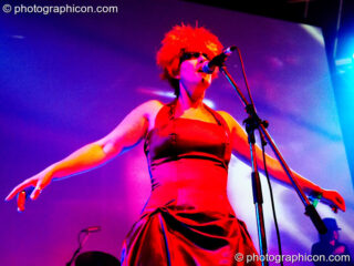 A woman in flame red dress, orange wig, and black painted eyes (like Pris from Blade Runner) is washed in blue light while performing with Juno Reactor on the Glade Stage at Glade Festival 2006. Aldermaston, United Kingdom. © 2006 Photographicon