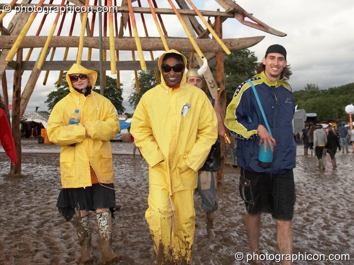 A small group of people clad in yellow pass underneath a large wooden decorative structure standing in the mud at Glade Festival 2007. Aldermaston, United Kingdom. © 2007 Photographicon