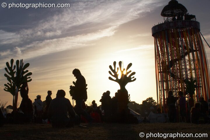 Silhouetted against the setting sun, a tall decorated scaffold tower with viewing platform rises above The Park next to cut-out sculptures of organic shapes at Glastonbury Festival 2008. Pilton, United Kingdom. © 2008 Photographicon