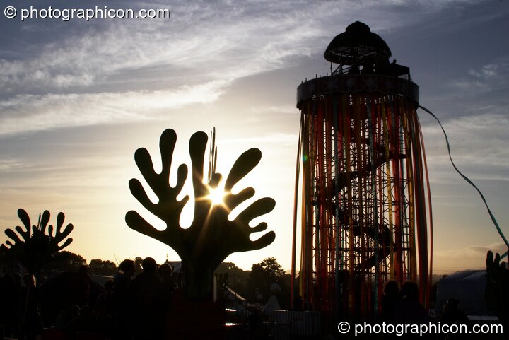 Silhouetted against the setting sun, a tall decorated scaffold tower with viewing platform rises above The Park next to cut-out sculptures of organic shapes at Glastonbury Festival 2008. Pilton, United Kingdom. © 2008 Photographicon