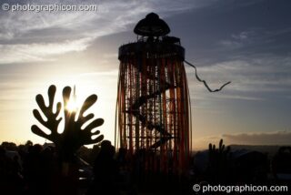 Silhouetted against the setting sun, a tall decorated scaffold tower with viewing platform rises above The Park next to cut-out sculptures of organic shapes at Glastonbury Festival 2008. Pilton, United Kingdom. © 2008 Photographicon