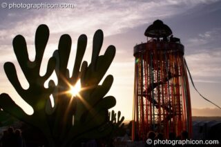 Silhouetted against the setting sun, a tall decorated scaffold tower with viewing platform rises above The Park next to cut-out sculptures of organic shapes at Glastonbury Festival 2008. Pilton, United Kingdom. © 2008 Photographicon