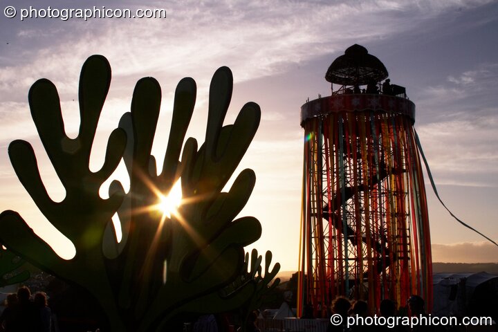 Silhouetted against the setting sun, a tall decorated scaffold tower with viewing platform rises above The Park next to cut-out sculptures of organic shapes at Glastonbury Festival 2008. Pilton, United Kingdom. © 2008 Photographicon