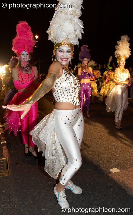 Woman with while face, gold jewellery and tall layer-hat smiles during the carnival at the Thames Festival 2005. London, United Kingdom. © 2005 Photographicon