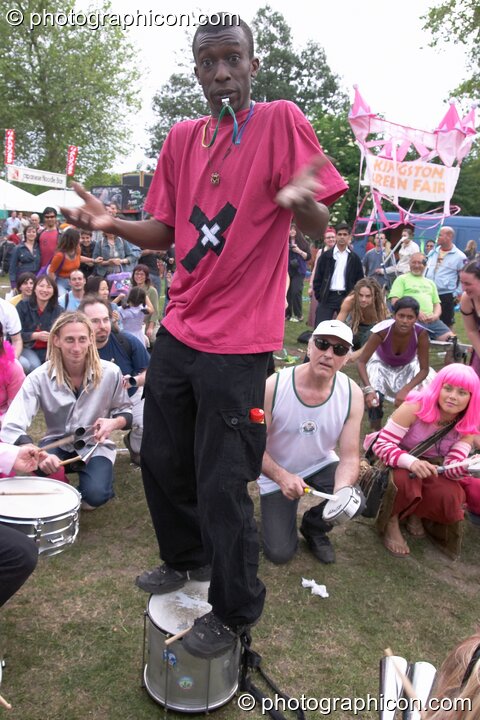 The leader of Rhythms of Resistance stands on his drum and shrugs at Kingston Green Fair 2004. Kingston Upon Thames, United Kingdom. © 2004 Photographicon