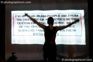 Woman stands with her arms raised in a cross silhouetted against projected agitprop text at The Synergy Project. London, United Kingdom. © 2005 Photographicon