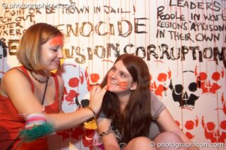 A woman paints another's face sitting next to an urban art painting by Genny & Ron in the Future Music room at Luminopolis (formerly The Synergy Project). London, United Kingdom. © 2008 Photographicon
